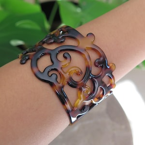 Può includere: Un bracciale a polsino largo e traforato con motivo tartarugato. Il bracciale presenta un motivo a vortice ripetuto nelle tonalità del marrone, nero e ambra. Il gioiello è indossato sul braccio di una persona.