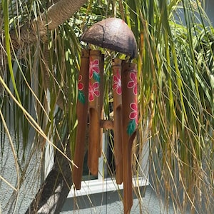 May include: A handcrafted wind chime made from bamboo and a coconut shell. The brown bamboo tubes are adorned with hand-painted red flowers and green leaves. The wind chime hangs from a tree branch, with green foliage in the background.