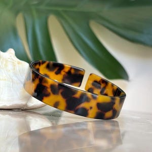 May include: A tortoiseshell-patterned cuff bracelet. The open-ended bangle features a warm palette of brown and black hues. The bracelet is displayed on a reflective surface, with a seashell and green leaf in the background.