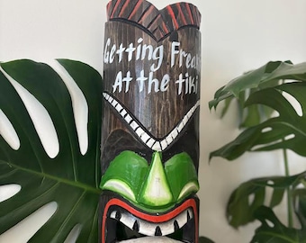 May include: A carved wooden tiki statue with a green face, red mouth, and white teeth. The statue has the text "Getting Freaky At the tiki" in white. The top of the statue is brown with red accents.