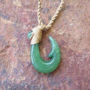 May include: A green jade fish hook pendant necklace with a gold wire wrap. The necklace is on a brown rope chain.