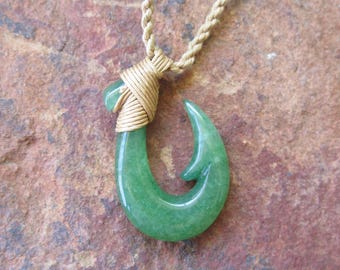 Puede incluir: Un collar con un colgante de anzuelo de pescado de jade verde y una envoltura de alambre dorado. El collar está en una cadena de cuerda marrón.