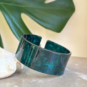 May include: A wide, open-cuff bracelet in a marbled, dark green pattern. The bracelet is made of a glossy material and has a rectangular shape with rounded edges. A large green leaf and a seashell are in the background.