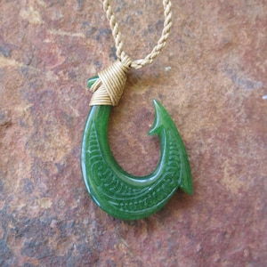 Puede incluir: Un collar con un colgante de anzuelo de pescado de jade verde y un cordón dorado. El anzuelo está tallado con diseños intrincados.