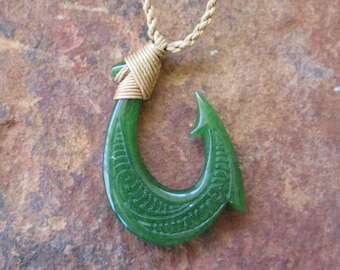 Puede incluir: Un collar con un colgante de anzuelo de pescado de jade verde y un cordón dorado. El anzuelo está tallado con diseños intrincados.