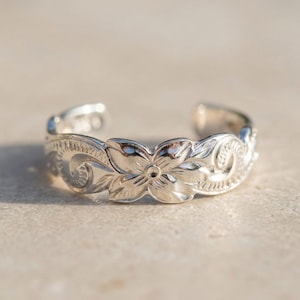 May include: A silver-colored toe ring with an open design. The ring features a floral pattern with detailed carvings of flowers and swirling leaves. The ring is displayed on a neutral-toned surface, highlighting the intricate design.