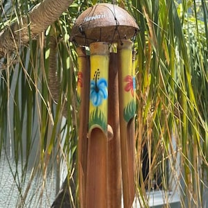 May include: A handcrafted wind chime with a coconut shell top and bamboo tubes. The tubes are painted yellow with floral designs and the word "Hawaii" is on the top. The wind chime hangs from a black string.