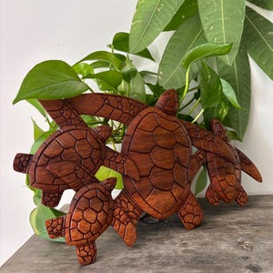 Könnte beinhalten: Eine Wandskulptur aus Holz mit vier Meeresschildkröten, die in einer rotbraunen Farbe geschnitzt sind. Die Schildkröten sind in einer geschichteten Formation angeordnet, wobei die größte Schildkröte hinten und die kleinste vorne ist.