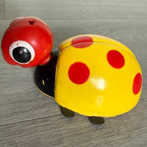 May include: A colorful toy ladybug with a red head, a yellow body with red spots, and black accents. The ladybug has large, cartoonish eyes and small, black feet. The toy is made of plastic and appears to be vintage.