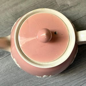 Op de afbeelding: Een roze keramische theepot met een bijpassend deksel en een witte handgreep en tuit. De theepot heeft een ronde body en een decoratieve witte rand rond de basis en het deksel. Het deksel heeft een kleine knop.