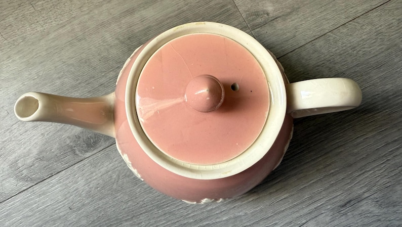 Op de afbeelding: Een roze keramische theepot met een bijpassend deksel en een witte handgreep en tuit. De theepot heeft een ronde body en een decoratieve witte rand rond de basis en het deksel. Het deksel heeft een kleine knop.