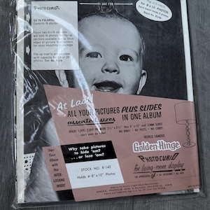 Puede incluir: Un paquete de álbum de fotos vintage con una imagen en blanco y negro de un bebé sonriente. El embalaje incluye texto sobre el almacenamiento de fotos, la capacidad del álbum y la marca "Golden-Hinge". El álbum contiene fotos de 10,16 cm x 20,32 cm y 20,32 cm x 25,4 cm.