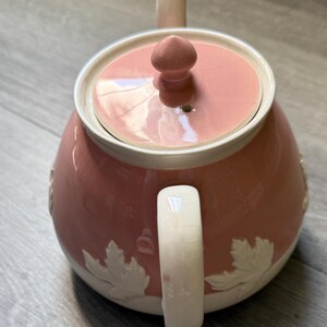 Op de afbeelding: Een roze keramische theepot met bijpassend deksel en handvat. De theepot heeft witte bladeren rond de basis en een witte rand. Het deksel heeft een kleine knop voor eenvoudig verwijderen. De tuit en het handvat zijn een lichtere roze tint.