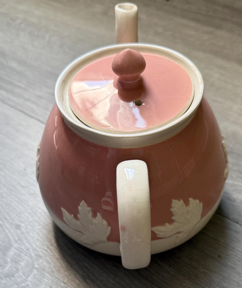 Op de afbeelding: Een roze keramische theepot met bijpassend deksel en handvat. De theepot heeft witte bladeren rond de basis en een witte rand. Het deksel heeft een kleine knop voor eenvoudig verwijderen. De tuit en het handvat zijn een lichtere roze tint.