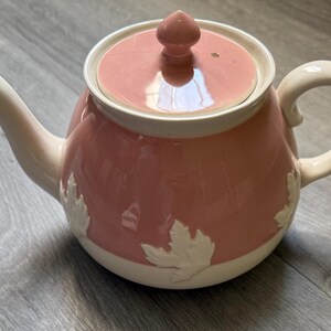 Op de afbeelding: Roze keramische theepot met een witte rand, handvat en tuit. De theepot heeft een deksel met een kleine knop en witte bladeren op de body. Een klassieker voor het serveren van thee.