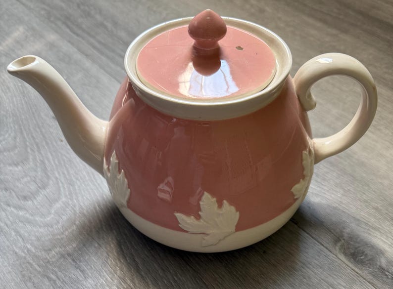 Op de afbeelding: Roze keramische theepot met een witte rand, handvat en tuit. De theepot heeft een deksel met een kleine knop en witte bladeren op de body. Een klassieker voor het serveren van thee.