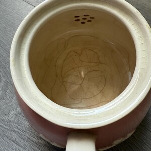 Op de afbeelding: Een vintage, tweekleurige keramische theepot met een cr&egrave;mekleurige rand en handvat en een roze body. De binnenkant is lichtbruin van kleur. De theepot heeft een kleine tuit en een reeks kleine gaatjes aan de bovenkant.