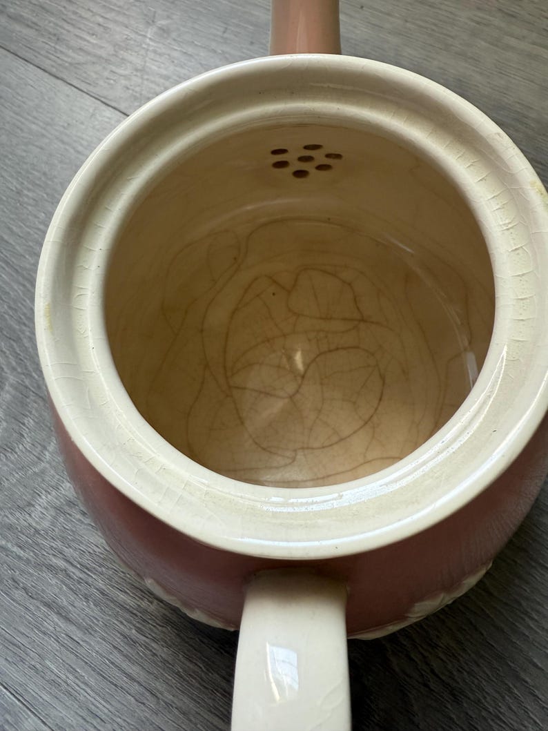 Op de afbeelding: Een vintage, tweekleurige keramische theepot met een cr&egrave;mekleurige rand en handvat en een roze body. De binnenkant is lichtbruin van kleur. De theepot heeft een kleine tuit en een reeks kleine gaatjes aan de bovenkant.