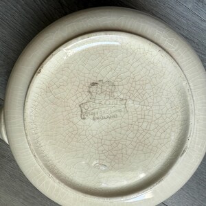 Op de afbeelding: Cr&egrave;me-kleurige keramische serveerschaal met een craquel&eacute; glazuur. De onderkant toont een vervaagd embleem en de tekst "Staffordshire England". De schaal heeft een ronde vorm, een opstaande rand en twee kleine handvatten.