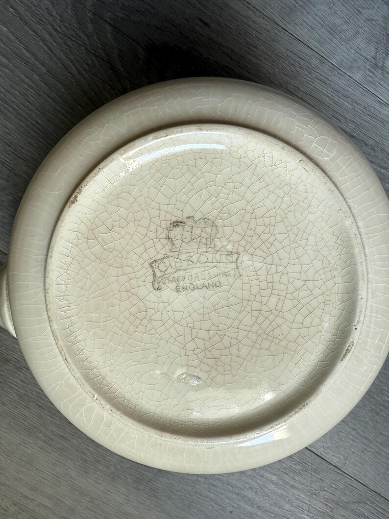Op de afbeelding: Cr&egrave;me-kleurige keramische serveerschaal met een craquel&eacute; glazuur. De onderkant toont een vervaagd embleem en de tekst "Staffordshire England". De schaal heeft een ronde vorm, een opstaande rand en twee kleine handvatten.