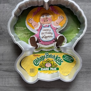 Puede incluir: Un molde para pastel de Cabbage Patch Kids de color plateado. El molde presenta un diseño de un Cabbage Patch Kid con cabello rubio y un vestido rosa. Las palabras "Happy Birthday Lauren" están escritas en el pastel. El molde es de Wilton.