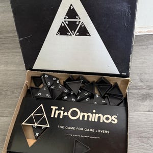 May include: Tri-Ominos board game box, open to reveal black triangular game pieces with white numbers. The box is black with white text and a triangle design. The game is by Pressman.