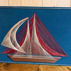 Puede incluir: Un barco de vela de arte de hilo con velas de hilo rojo, blanco y beige y un casco de madera marrón, sobre un fondo azul. La obra de arte está enmarcada con un borde de metal plateado, creando un tema náutico.