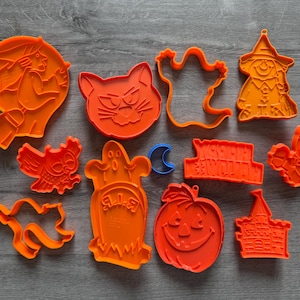 May include: A collection of orange Halloween-themed cookie cutters in various shapes, including a witch on a broom, a cat, a ghost, an owl, a pumpkin, a tombstone, a haunted house, and a bat. A blue crescent moon is also included.