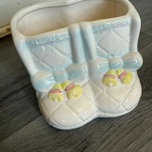 Könnte beinhalten: Ein Keramik-Pflanzgefäß in Form eines Kinderwagens. Der Pflanztopf ist weiß mit hellblauen Akzenten und gelb-rosa floralen Details. Der Wagen hat ein Rautenmuster und eine rechteckige Öffnung.