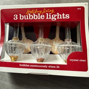 Könnte beinhalten: Eine Schachtel mit 3 Holiday Living Blasenlichtern. Die Schachtel ist rot und weiß mit den Worten "3 bubble lights" in Weiß. Die Lichter sind kristallklar mit goldenen und kupferfarbenen Akzenten. Der Text "bubbles continuously when lit" ist ebenfalls sichtbar.