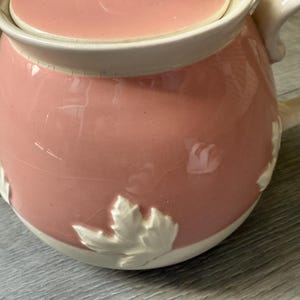 Op de afbeelding: Een roze keramische theepot met bijpassende deksel. De theepot heeft een witte rand en handvat. Witte bladeren zijn in reli&euml;f op het roze lichaam. De theepot staat op een grijze ondergrond.
