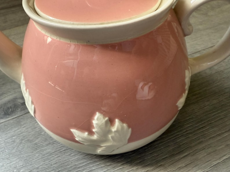 Op de afbeelding: Een roze keramische theepot met bijpassende deksel. De theepot heeft een witte rand en handvat. Witte bladeren zijn in reli&euml;f op het roze lichaam. De theepot staat op een grijze ondergrond.