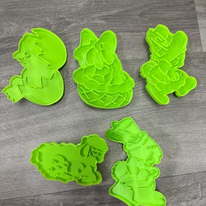 Puede incluir: Cinco cortadores de galletas de Pascua de color verde brillante. Las formas incluyen un pollito saliendo de un huevo, un conejito en una cesta, un duende, una oveja y un segundo duende. Los cortadores son de plástico y están sobre una superficie de madera gris.