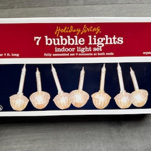 Puede incluir: Una caja de 7 luces de burbujas Holiday Living, un juego de luces para interiores. La caja tiene una parte superior roja con texto blanco y una parte inferior azul oscuro con siete luces de burbujas transparentes. El texto dice "más de 2,13 m de largo" y "cristalino".
