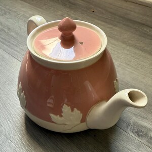 Op de afbeelding: Een roze keramische theepot met een cr&egrave;mekleurige handgreep, tuit en dekselrand. Het deksel heeft een kleine knop en een gat. Witte bladeren zijn in reli&euml;f op het lichaam van de theepot.