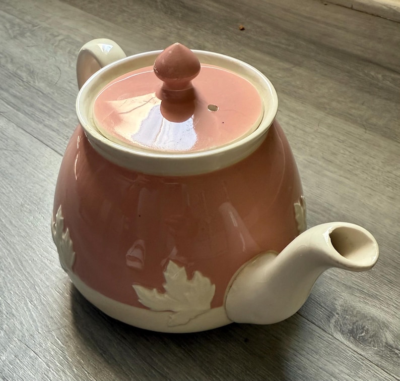 Op de afbeelding: Een roze keramische theepot met een cr&egrave;mekleurige handgreep, tuit en dekselrand. Het deksel heeft een kleine knop en een gat. Witte bladeren zijn in reli&euml;f op het lichaam van de theepot.