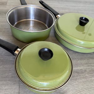 May include: A set of three sage green cooking pans with black handles and matching lids. One pan is shown without a lid, revealing a stainless steel interior. Each lid features a black knob handle. The exterior of the pans is a smooth, solid sage green.