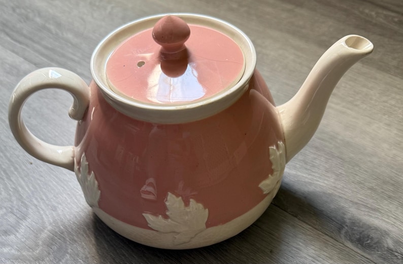 Op de afbeelding: Een roze en witte keramische theepot met deksel en handvat. De theepot heeft een afgeronde vorm met witte bladeren bij de basis. Het handvat en de tuit zijn wit. Het deksel heeft een kleine knop.