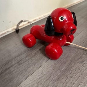 Può includere: Un giocattolo a forma di cane rosso in legno da tirare, con orecchie nere e un collare nero. Il cane ha una coda a molla e una corda per tirare. Il giocattolo è su un pavimento di legno grigio.