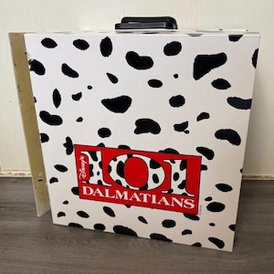 Könnte beinhalten: Eine weiße Box mit schwarzen Dalmatinerflecken und den Worten "101 Dalmatians" in einem roten Rechteck. Die Box hat einen schwarzen Griff und ist wahrscheinlich ein Aufbewahrungsbehälter oder ein dekorativer Gegenstand.