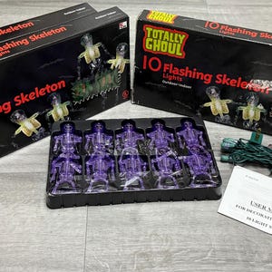 May include: Several black boxes with the text "Totally Ghoul 10 Flashing Skeleton Lights" are shown. Inside, purple skeleton lights are visible. A user manual and a string of green lights are also present, suggesting Halloween decorations.
