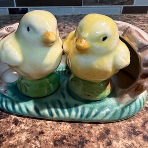 May include: A ceramic figurine featuring two yellow chicks perched on green bases, with a brown and white eggshell-shaped accent. The base is a light green with a textured design. The chicks have black eyes and orange beaks.