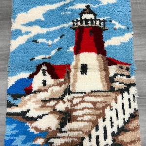 May include: A handmade, textured wall hanging depicting a lighthouse scene. The lighthouse features a red tower and a white base, set against a blue sky with white clouds. The artwork includes a rocky shoreline and a blue ocean.
