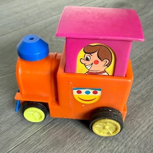 May include: A vintage toy train in bright orange, with a pink roof and blue accents. The train features a smiling cartoon face and yellow wheels. A classic toy for children.