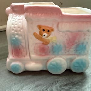 Könnte beinhalten: Ein keramik-zugförmiger Pflanztopf in Pastellrosa, Weiß und Blau. Der Pflanztopf hat ein Teddybär-Design und florale Akzente. Die Räder und floralen Akzente sind blau. Der Teddybär ist hellbraun.