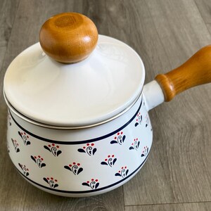 Puede incluir: Cacerola de esmalte blanco con asa y tapa de madera. La olla presenta un estampado floral en azul marino y rojo. La tapa tiene un pomo de madera. El asa y el pomo son de madera clara.