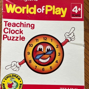 May include: A red, yellow, and blue box for a children's clock puzzle game. The box features a smiling cartoon clock face with sunglasses and the text "Waddingtons World of Play Teaching Clock Puzzle 4+ YEARS". The bottom of the box says "This Game Teaches TELLING TIME" and has a logo with a turtle and the text "Children Are Our Future".