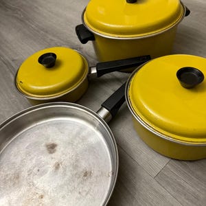 以下が含まれることがあります： 黄色いエナメル加工が施された調理器具3点セット。蓋付きの鍋2つと、蓋のないフライパン1つが含まれています。各鍋には黒いハンドルと蓋に黒いノブが付いています。フライパンは銀色の内面と黒いハンドルを備えています。