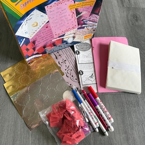 May include: A Crayola My Style Stationery Design kit with various stationery supplies. The kit includes a box, markers, stickers, stencils, pink paper, and a pad of white paper. The box is yellow and green with the words "Crayola" and "Express Yourself".