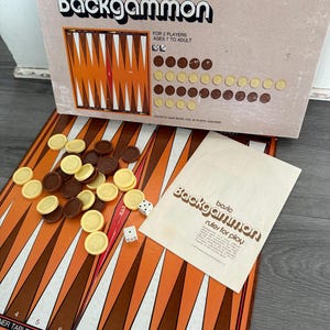 Puede incluir: Un juego de Backgammon vintage con un tablero naranja y marrón, fichas marrones y amarillas, dados y un libro de reglas. La caja dice "Backgammon" y "Para 2 jugadores, a partir de 7 años."